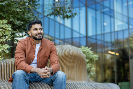 Young indian man with a beard and wearing casual clothes sits on a modern bench in an urban setting,...