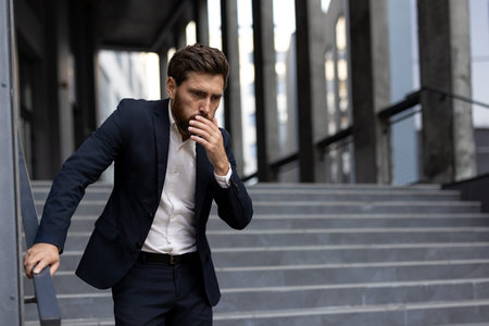 Businessman suffering from nausea covering his mouth with hand while standing on stairs and feeling unwell, possibly due to food poisoning, virus infection or other illnessの写真素材