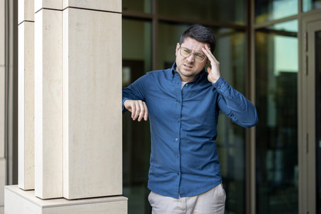 A man in glasses and casual clothing, showing signs of stress near an urban structure.