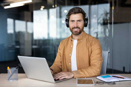 Young smiling business man with beard wearing headphones, typing on a laptop, enjoying remote work...