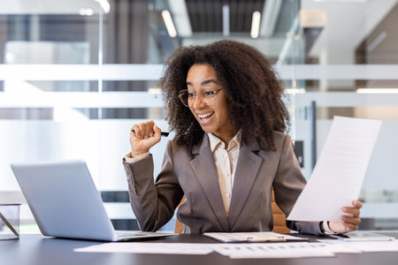 Happy young African American woman sitting in the office at the desk, holding documents in her hands, looking at the laptop screen and enjoying the news and success.の写真素材