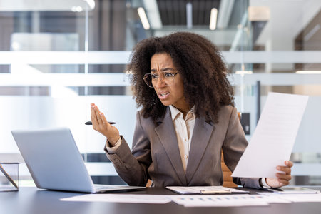 Angry and upset young african american business woman sitting in office at desk, holding documents and gesturing unhappily at laptop screen.の写真素材