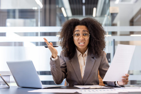 Portrait of angry and disappointed African American woman sitting in office at workplace, holding papers and gesturing emotionally to camera.の写真素材