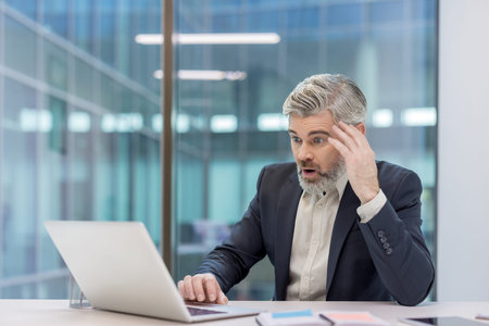 Mature businessman experiencing shock and disbelief while looking at a laptop computer screen,...