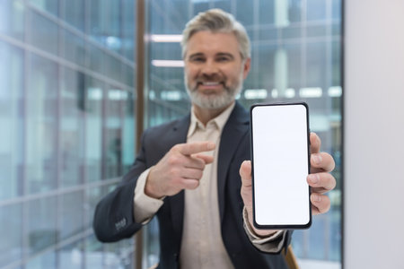 Senior businessman in a suit smiling and pointing at a smartphone with a blank white screen in a modern office, presenting a mobile app mockup for business promotionの写真素材