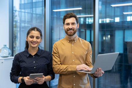 Two smiling business colleagues are standing in a modern office, using a digital tablet and laptop, showcasing teamwork and connectivity in the workplaceの写真素材