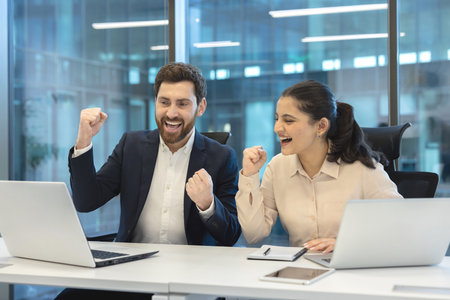 Two colleagues celebrate a business win, cheering and pumping fists while working on laptops at a modern office desk, expressing teamwork, excitement, and professional achievementの写真素材
