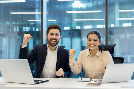 Two happy business colleagues are raising their fists in a modern office, expressing their...
