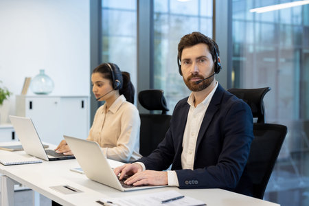 Two customer service representatives collaborating in a bright, modern office, using laptops and...