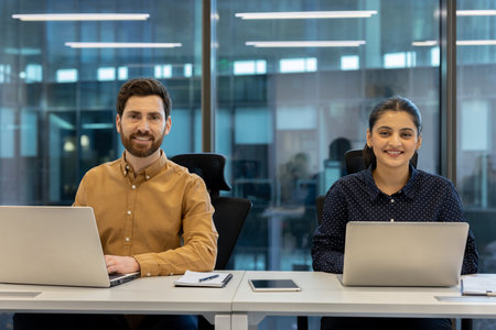 Two smiling business colleagues sitting at a desk in a modern office, working on laptops and enjoying a productive day filled with collaboration and teamworkの写真素材
