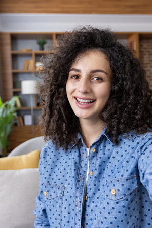 Young woman with curly hair smiling warmly at the camera during a video call at home, recording a...
