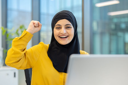 Muslim businesswoman wearing a hijab, cheering and showing a fist pump while working on a laptop in...