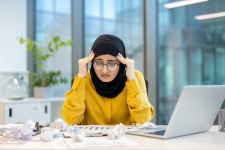 Young muslim businesswoman in hijab and glasses clutching her temples at a cluttered office desk, stressed and fatigued from overwork, missed deadlines and burnoutの写真素材