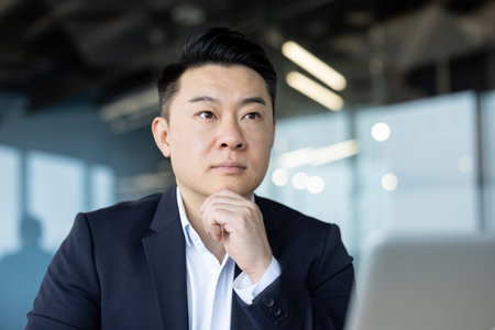 Photo of a serious young Asian man businessman in a suit sitting in the office at the workplace and thoughtfully looking ahead.の写真素材