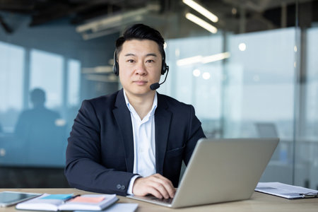 Portrait of a young Asian man wearing a headset and business suit sitting at a desk in the office, working on a laptop and looking confidently at the camera.の写真素材