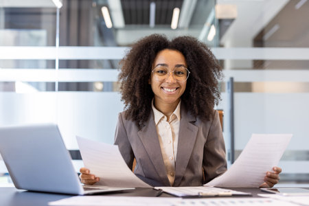 Portrait of a smiling young African American woman in a suit sitting at a desk in the office,...