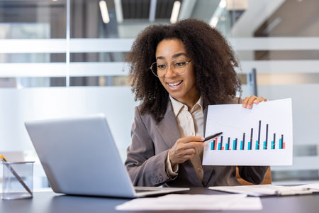 A smiling young African-American woman in a suit sits in the office at the table, conducts an online conference, shows documents with stickers on the laptop camera.の写真素材