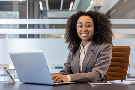 Portrait of a young smiling African American woman in a suit sitting in the office at a desk, using...