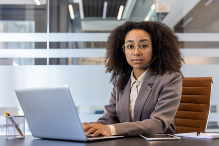 Serious and focused young African American woman in business suit and headset sitting in office at...