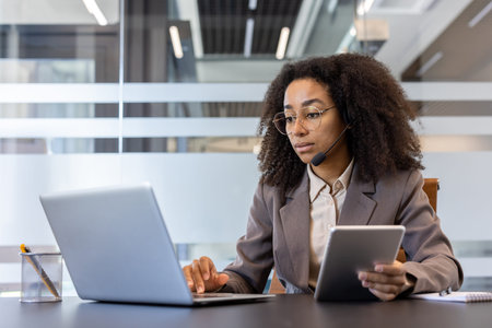 Serious and busy young African American woman wearing headset sitting at desk and working on laptop...