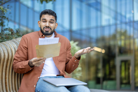 Young man sitting outdoors on a bench holding an open envelope with papers, looking confused and disappointed with the received news, reflecting on university failureの写真素材