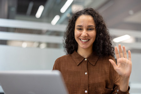 Young mixed-race woman smiling and waving at camera during an online video call on her laptop from a...