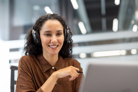 Woman wearing headphones smiles at laptop screen during an online meeting in a modern office,...