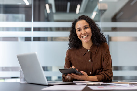 Professional businesswoman with curly hair smiling at her office desk, using a calculator and laptop...