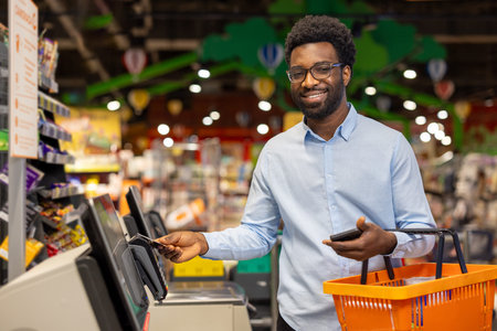Young african man smiling as he completes a contactless credit card payment with his smartphone at a...