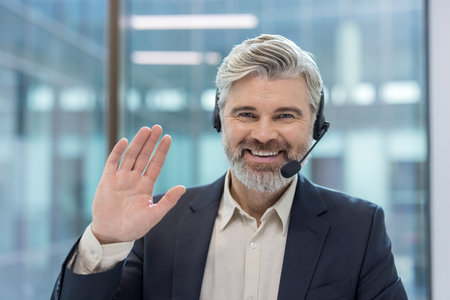 Friendly mature man working in customer service happily greeting while wearing a head-mounted microphone and headphones in a modern office environmentの写真素材