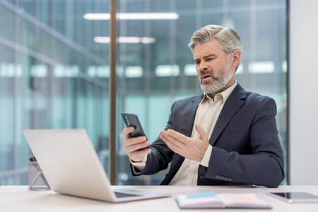 Senior businessman in suit at desk reacting with shock and frustration to bad news on his...