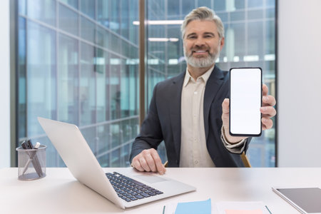 Mature businessman sitting at a modern office desk, holding a smartphone with a blank white screen up to the camera, presenting mobile technology and application displayの写真素材