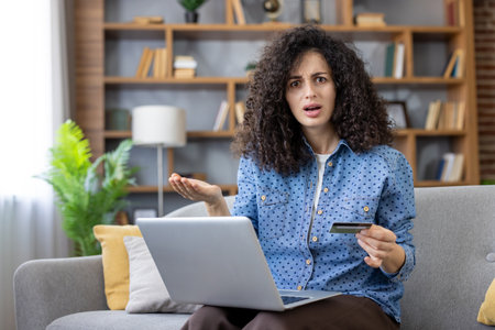 Woman feeling frustrated and confused, holding a credit card and looking at her laptop screen after encountering a problem with an online payment while shopping from her couchの写真素材