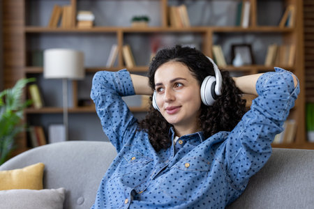 Smiling young woman with curly hair relaxing on a comfortable sofa at home, enjoying leisure time while listening to audio through wireless headphonesの写真素材