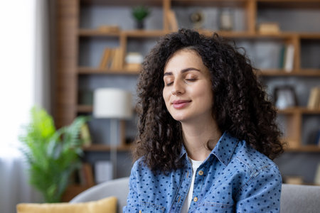 Woman with curly hair closing eyes meditating, practicing mindfulness, feeling peaceful and relaxed while sitting in the comfort of her living room, focusing on self-care and well-beingの写真素材