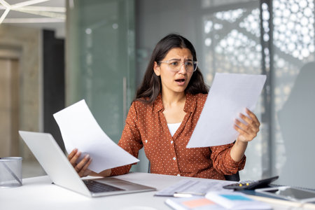 Young woman looking shocked and distressed, holding two documents in her hands, sitting at her office desk with a laptop and calculator, reacting to unexpected bad financial newsの写真素材