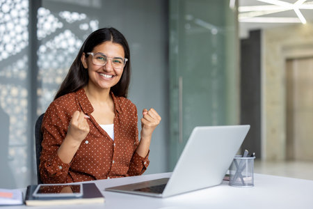 Businesswoman with glasses is sitting at her desk, raising her fists in a gesture of triumph, expressing her excitement and achievement after successfully completing a task on her laptopの写真素材