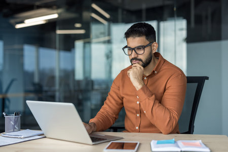 Indian businessman wearing glasses and an orange shirt concentrating and thinking while looking at a...