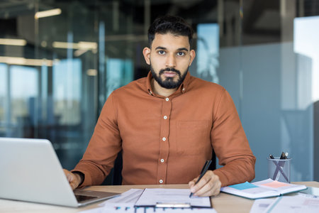 Confident indian businessman sitting at a desk, looking directly at the camera while working on a laptop, writing notes, and reviewing financial documents in a modern office setupの写真素材