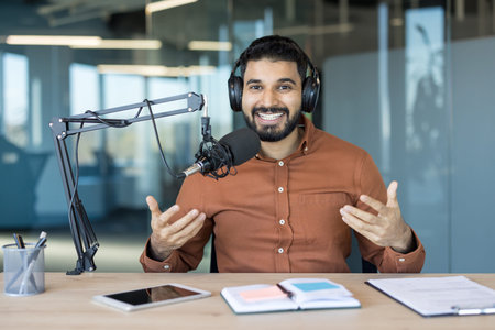 Smiling young man wearing headphones, recording a podcast with a microphone in a modern studio,...