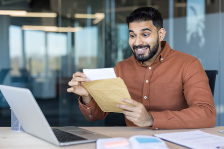 Excited young professional man sitting at his office desk opening a brown envelope with a surprised happy expression, learning about a promotion or financial successの写真素材