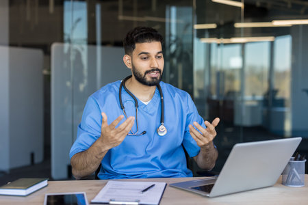 Indian doctor in blue scrubs and stethoscope having a video call with a patient on a laptop,...