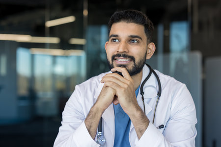Indian male doctor wearing a white lab coat and stethoscope, thoughtfully looking away, representing medical contemplation and professional aspiration in a clinic settingの写真素材