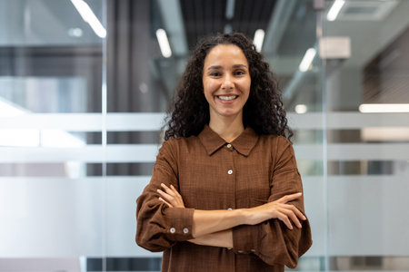 Professional businesswoman with curly hair smiling and looking at camera, standing with arms crossed in a modern corporate office, representing confidence and successの写真素材