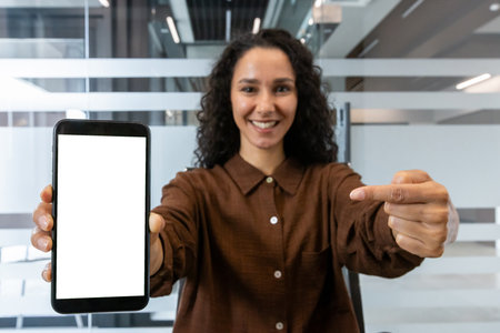 Young businesswoman smiling, holding a smartphone with a blank white screen, and pointing at it, showcasing technology in a contemporary office environmentの写真素材