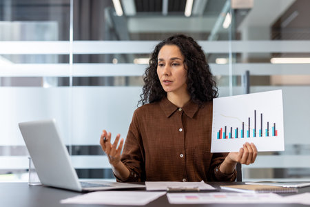 Businesswoman engaging in a video conference, presenting a bar chart and line graph on paper, discussing financial data, and collaborating remotely using her laptop in a modern officeの写真素材