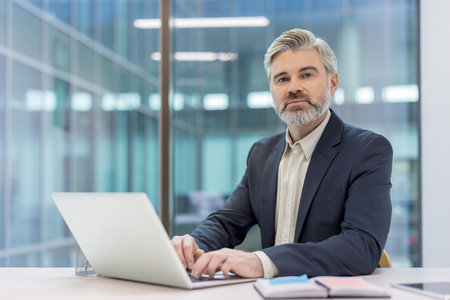 Confident mature businessman actively typing on a laptop, working on a project from a modern office, representing professionalism, success, and digital communication in a corporate environmentの写真素材