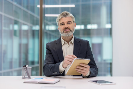 Mature executive businessman with a distinguished beard sitting at his office desk, looking at the camera and taking notes in a notebook during a professional video call or online conferenceの写真素材