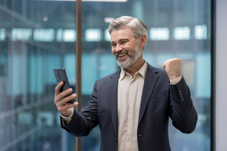 Mature businessman receiving good news on mobile phone, showing happiness and celebrating business...
