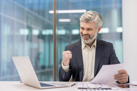 Mature businessman with gray hair and beard celebrating a corporate achievement, looking excited and happy while sitting at an office desk with a laptop and documentsの写真素材